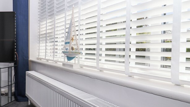 Une pièce avec un radiateur blanc, un téléviseur posé sur un meuble et une fenêtre avec des stores blancs.