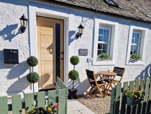 A white cottage with a wooden door, two windows, a small outdoor seating area with a table and chairs, and a sign reading "Lye Cottage." - Lyne Cottage, The Retreat (West Linton)