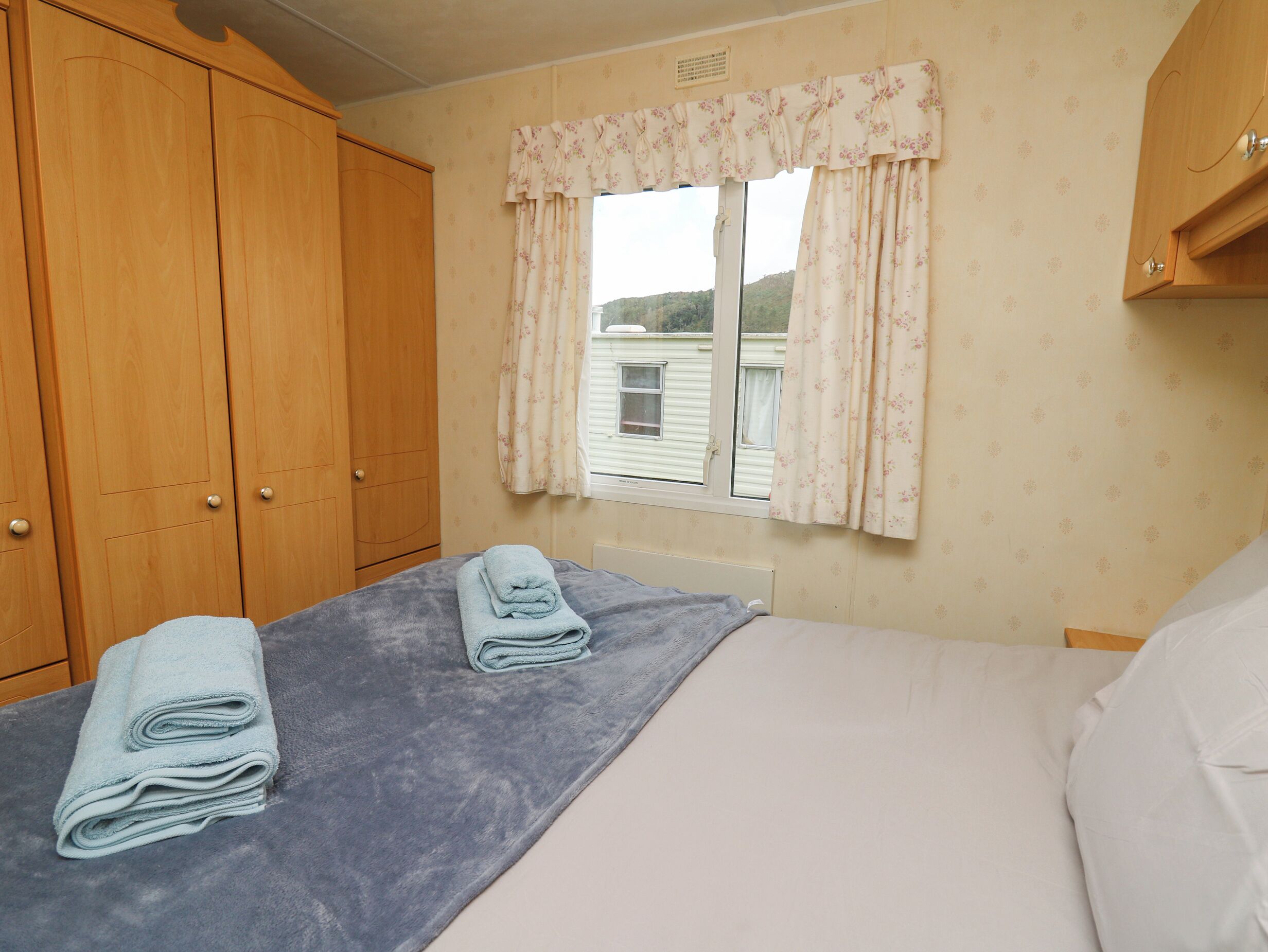 A bedroom with a bed, a window with floral curtains, and wooden wardrobes.