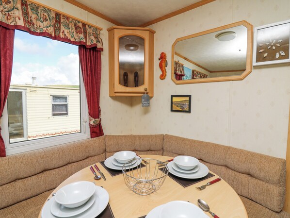A dining area with a round table set for four, surrounded by cushioned benches. A cabinet with glass doors is mounted on the wall, and a mirror hangs above the table. - D12 Treetops (Aberystwyth)