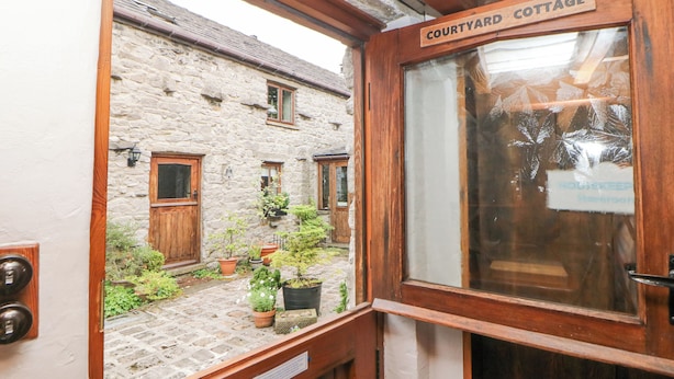 Une cour avec une porte en bois portant l’inscription « Maison de la cour », des murs en pierre et des plantes en pot.