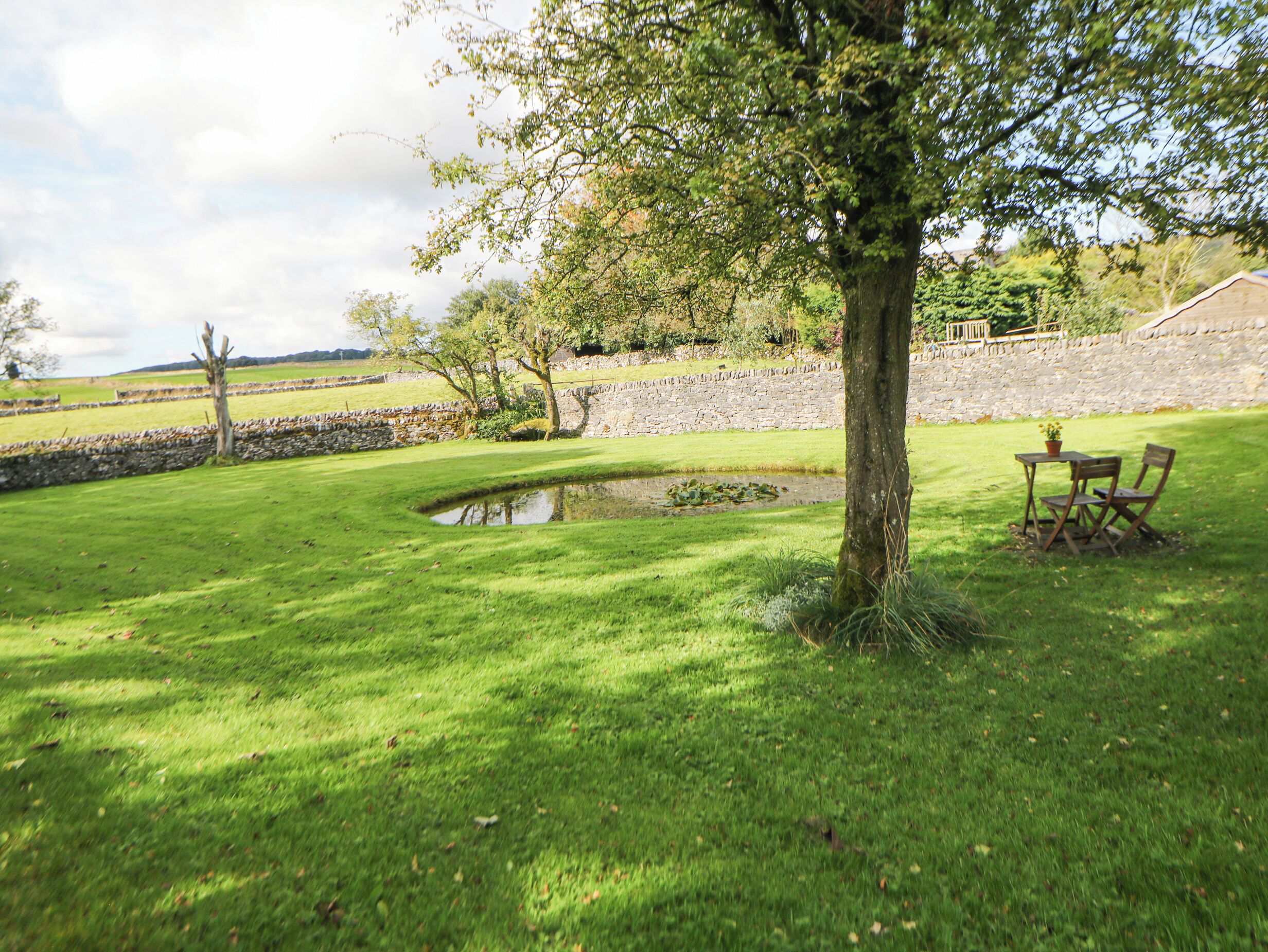 A grassy outdoor area with a pond, stone wall, and a table with chairs.