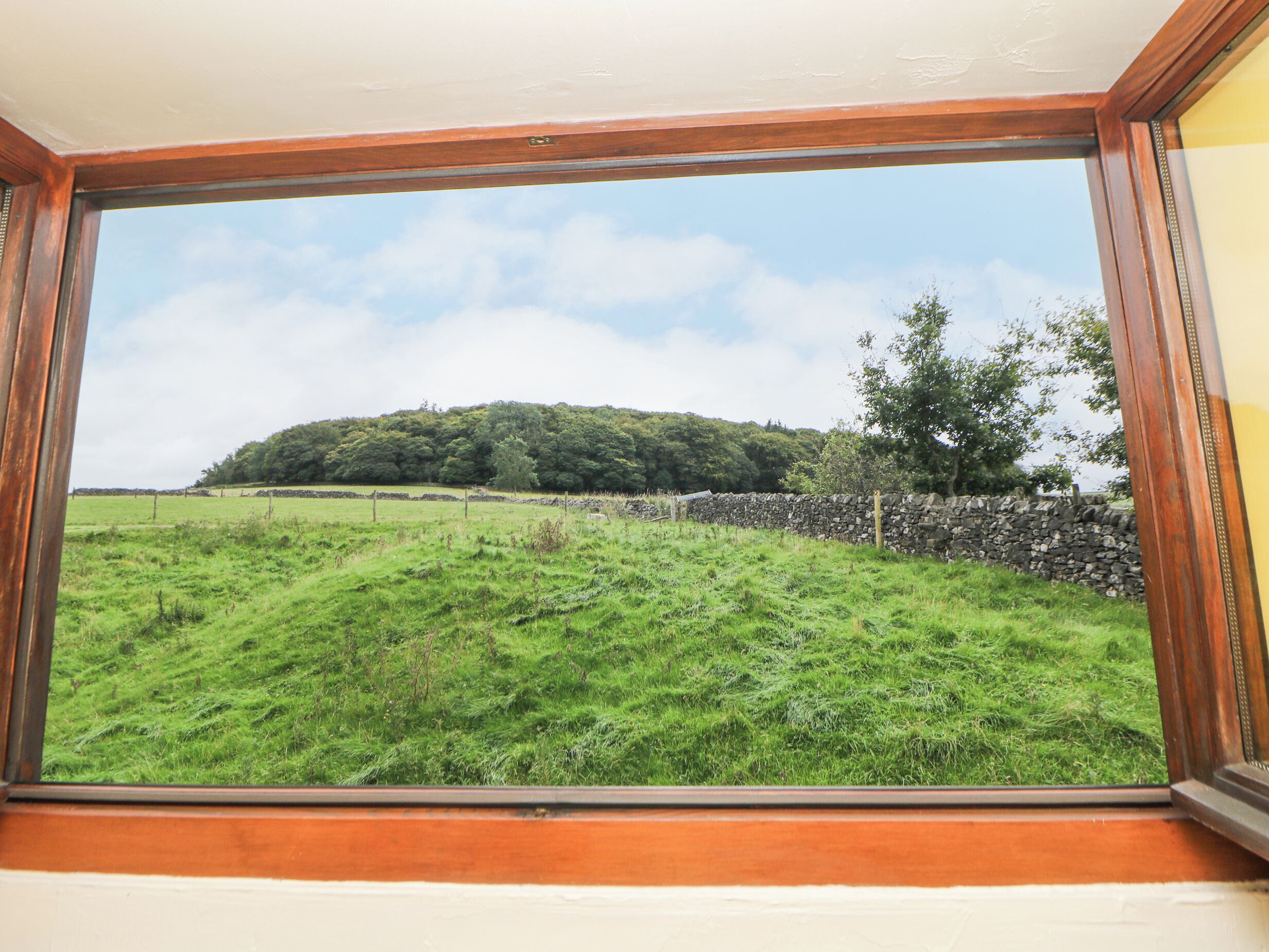Vista a través de una ventana de un campo cubierto de hierba, con un muro de piedra y una colina en la distancia.