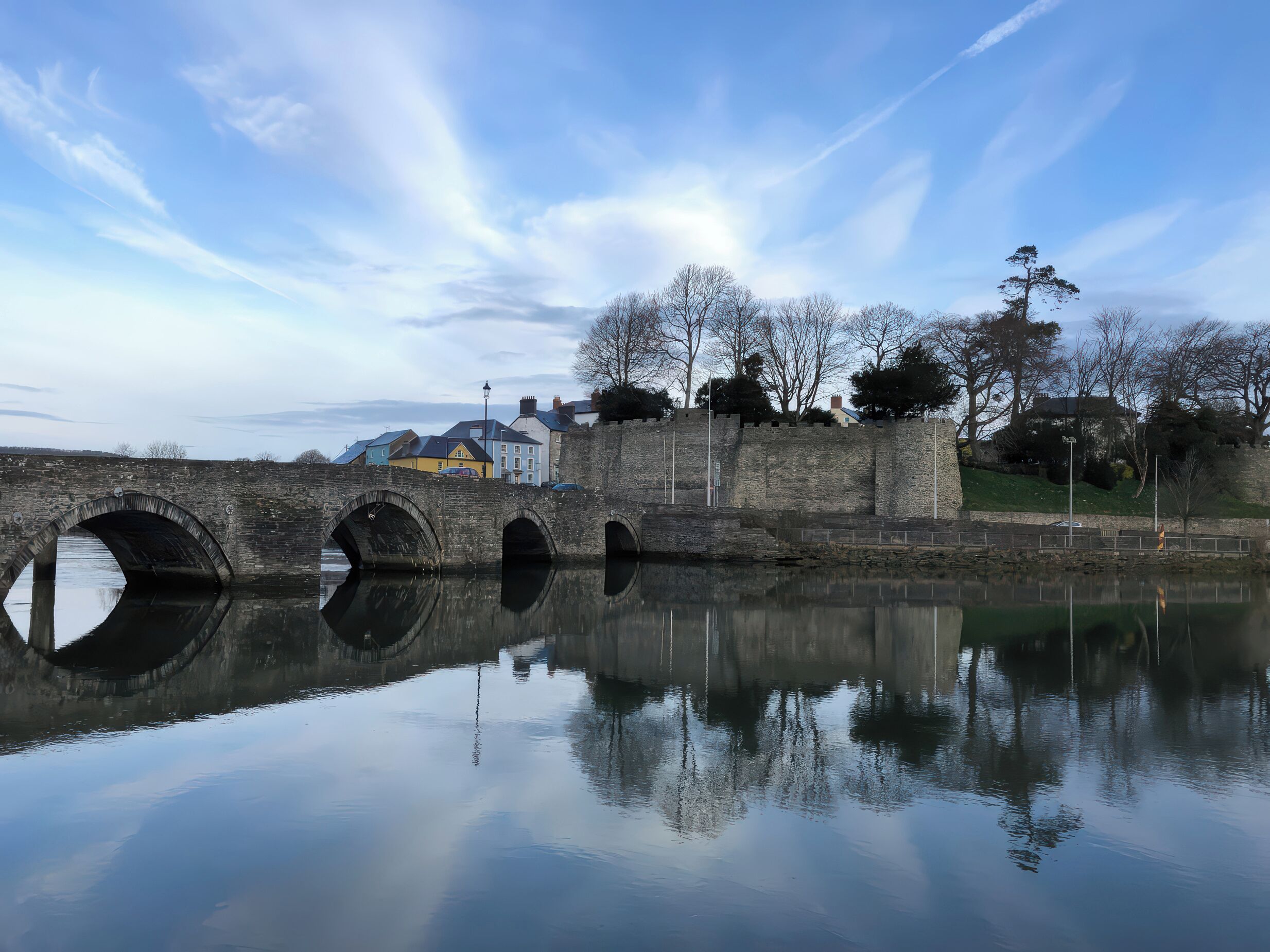 A stone bridge over a calm river with a clear reflection, surrounded by trees and buildings.