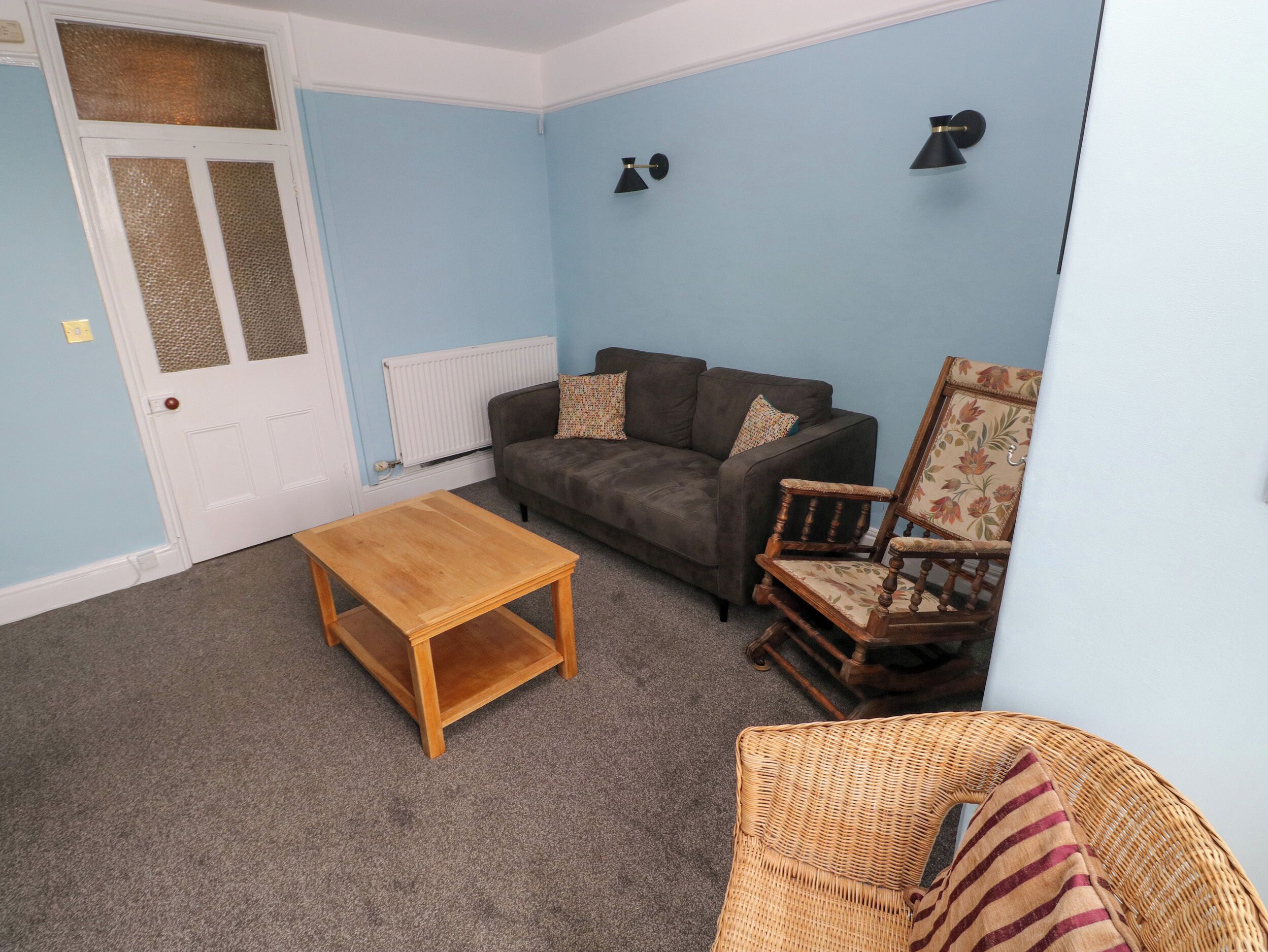 A living room with a sofa, a wooden coffee table, a wicker chair, and two wall-mounted lamps.