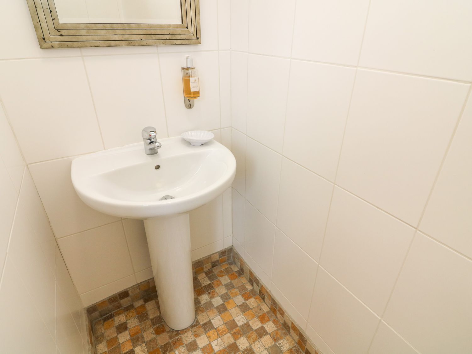 A bathroom with a white pedestal sink, a small mirror, and a soap dispenser. The floor has a checkered tile pattern.