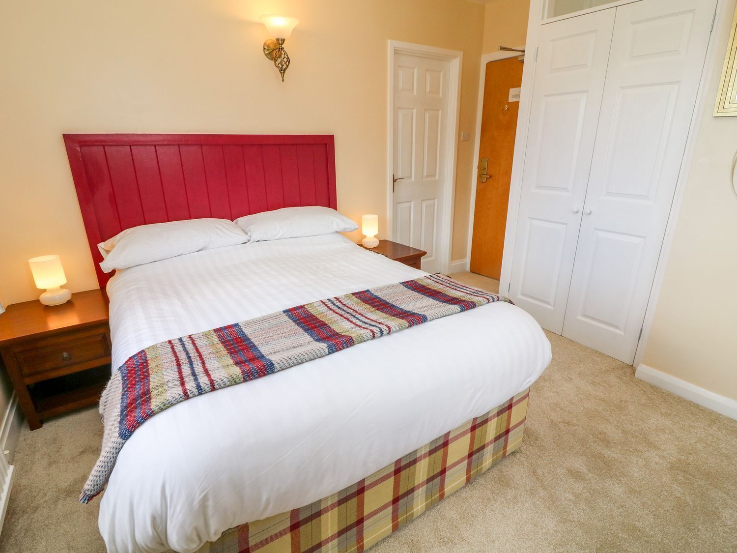A bedroom with a red headboard, white bedding, a colorful blanket, bedside lamps, and a carpeted floor.