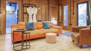 Interior de una cabaña de madera con un sofá, un sillón, un otoman redondo y una mesita.