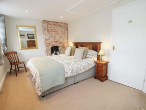 Een slaapkamer met een haard van baksteen, een bed met bloemenbedekking, een houten nachtkastje met een lamp, een stoel en een witte deur.