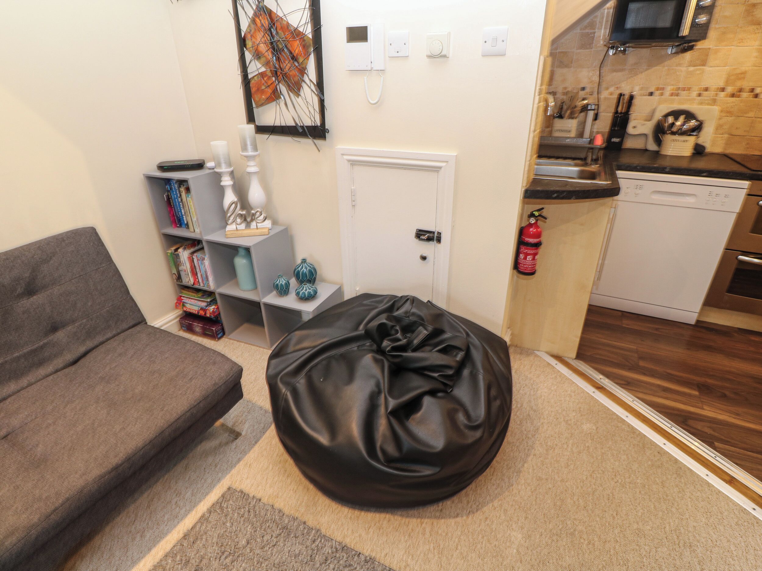 Um espaço compacto com um sofá cinzento, um pufe preto, uma estante com livros e objetos decorativos, uma porta branca e uma cozinha compacta com micro-ondas, pia e fogão.