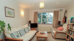 A cozy living room with a sofa, a coffee table, a bookshelf, a guitar, and a window with curtains. - Old Harry Cottage 16 (Swanage)
