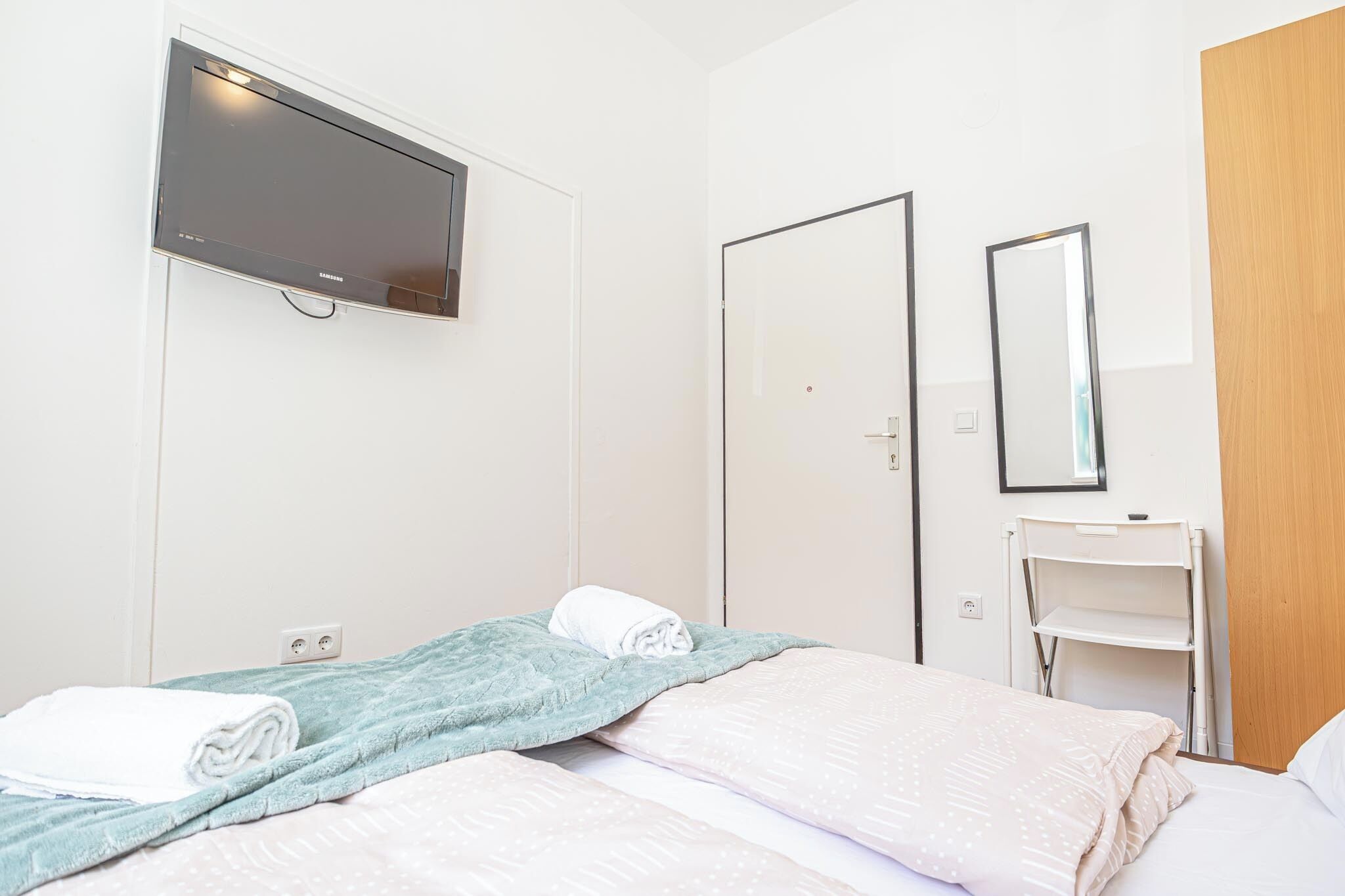 Une chambre d’hôtel avec un lit, un téléviseur fixé au mur, une porte, un miroir et une petite étagère blanche.