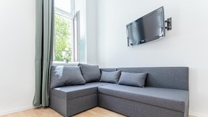 A modern living room with a grey sectional sofa, a mounted flat-screen TV, and a window with curtains. - 6BR Apt Lovely Garden Near Lorenz Böhler (Vienna)