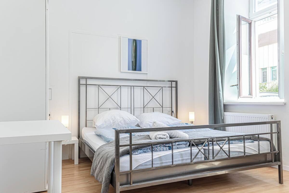A modern bedroom with a metal bed frame, a white bedside table, and a window with curtains.
