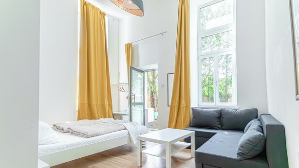 A modern, well-lit room with a bed, a sofa, a small table, and large windows with curtains. - 6BR Apt Lovely Garden Near Lorenz Böhler (Vienna)