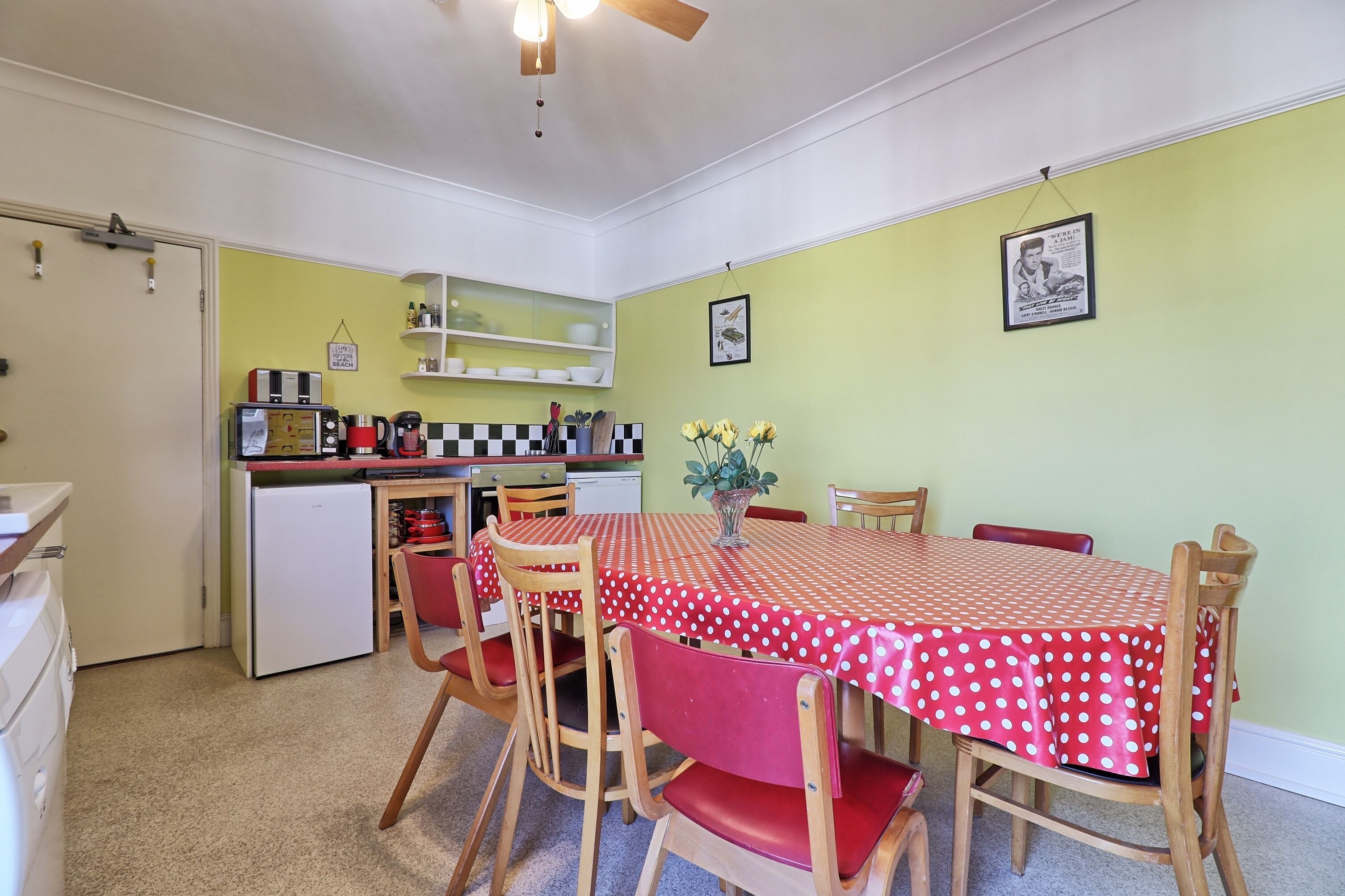 A compact kitchen with a refrigerator, microwave, and sink. There is a dining area with a table covered in a polka dot tablecloth and red chairs.