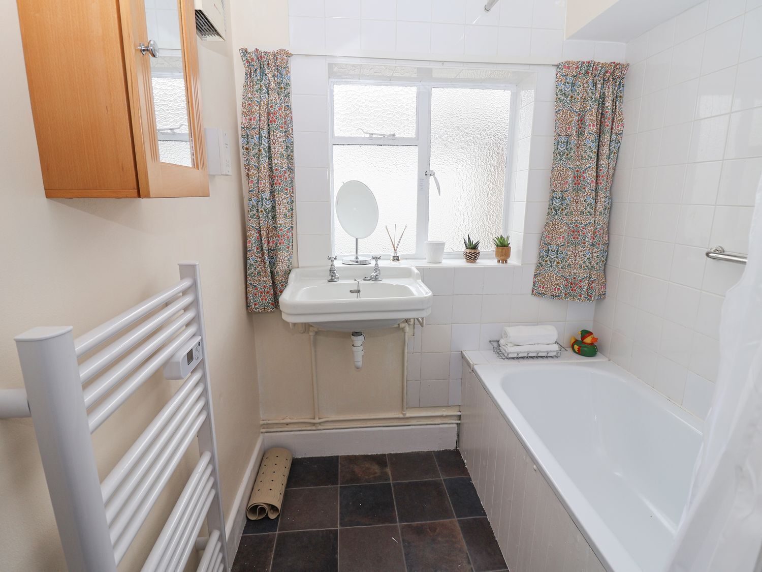 A compact bathroom with a white sink, mirror, and bathtub. There are floral curtains, a small window, and a radiator.