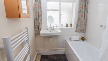 A compact bathroom with a white sink, mirror, and bathtub. There are floral curtains, a small window, and a radiator.
