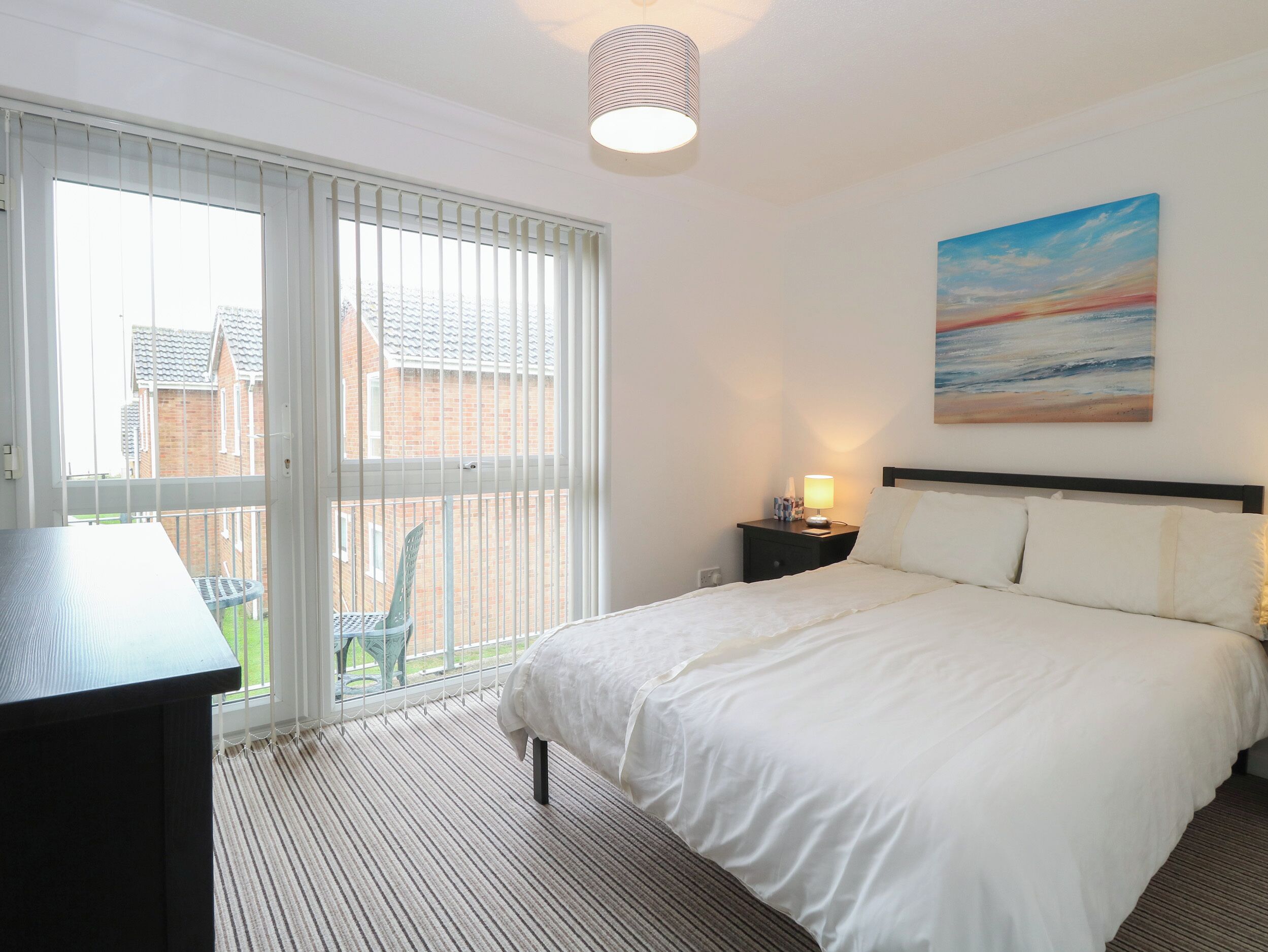 A hotel room with a bed, a desk, a chair, a painting, and a view of the outside.