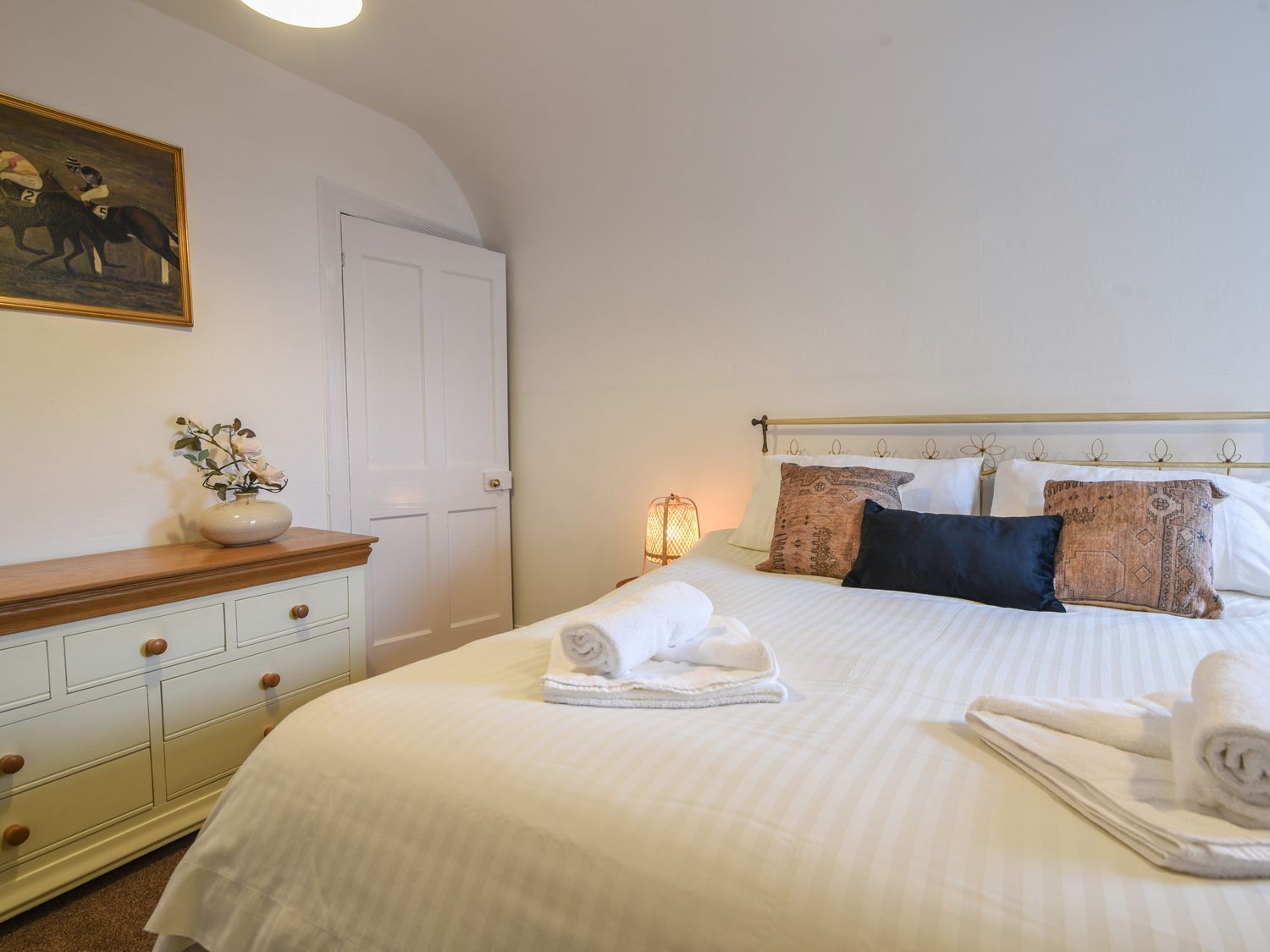 A neatly made bed with white linens and rolled towels, a wooden dresser with drawers, a wall-mounted painting of horses, and a white door.