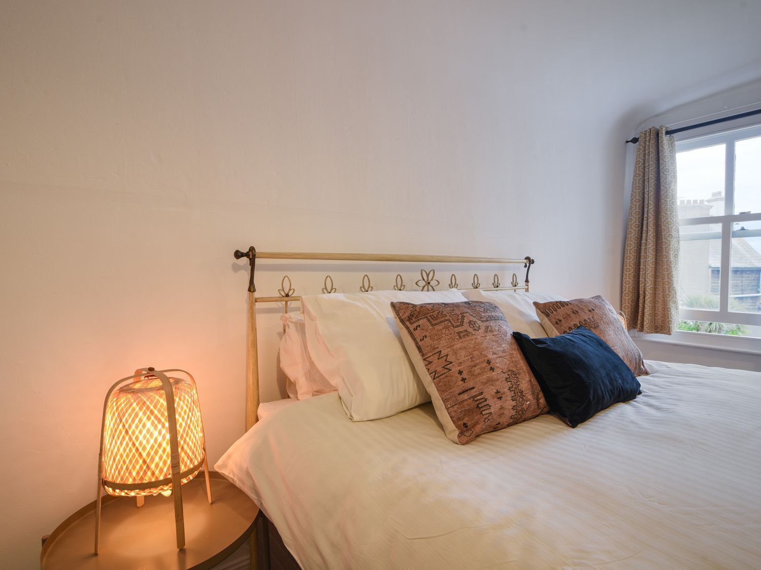 Una cama bien hecha con almohadones estampados, una lámpara de noche y una ventana con cortinas.