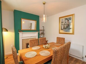 Un comedor con mesa de madera, sillas tejidas, chimenea blanca, un espejo y un cuadro en la pared.