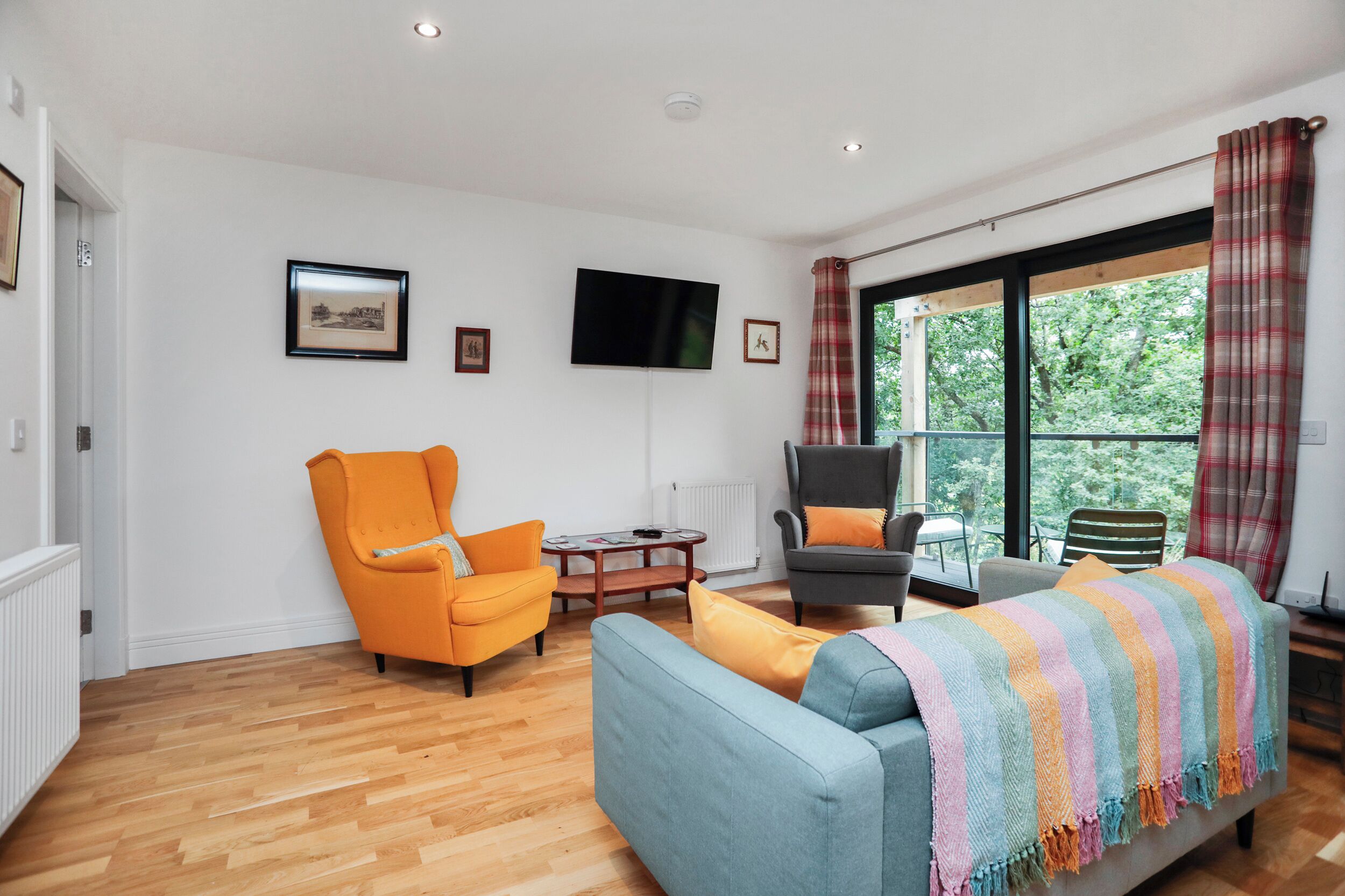 Un salon avec un canapé bleu, un fauteuil orange et un fauteuil gris. Il y a un téléviseur accroché au mur, une fenêtre avec des rideaux et un radiateur.