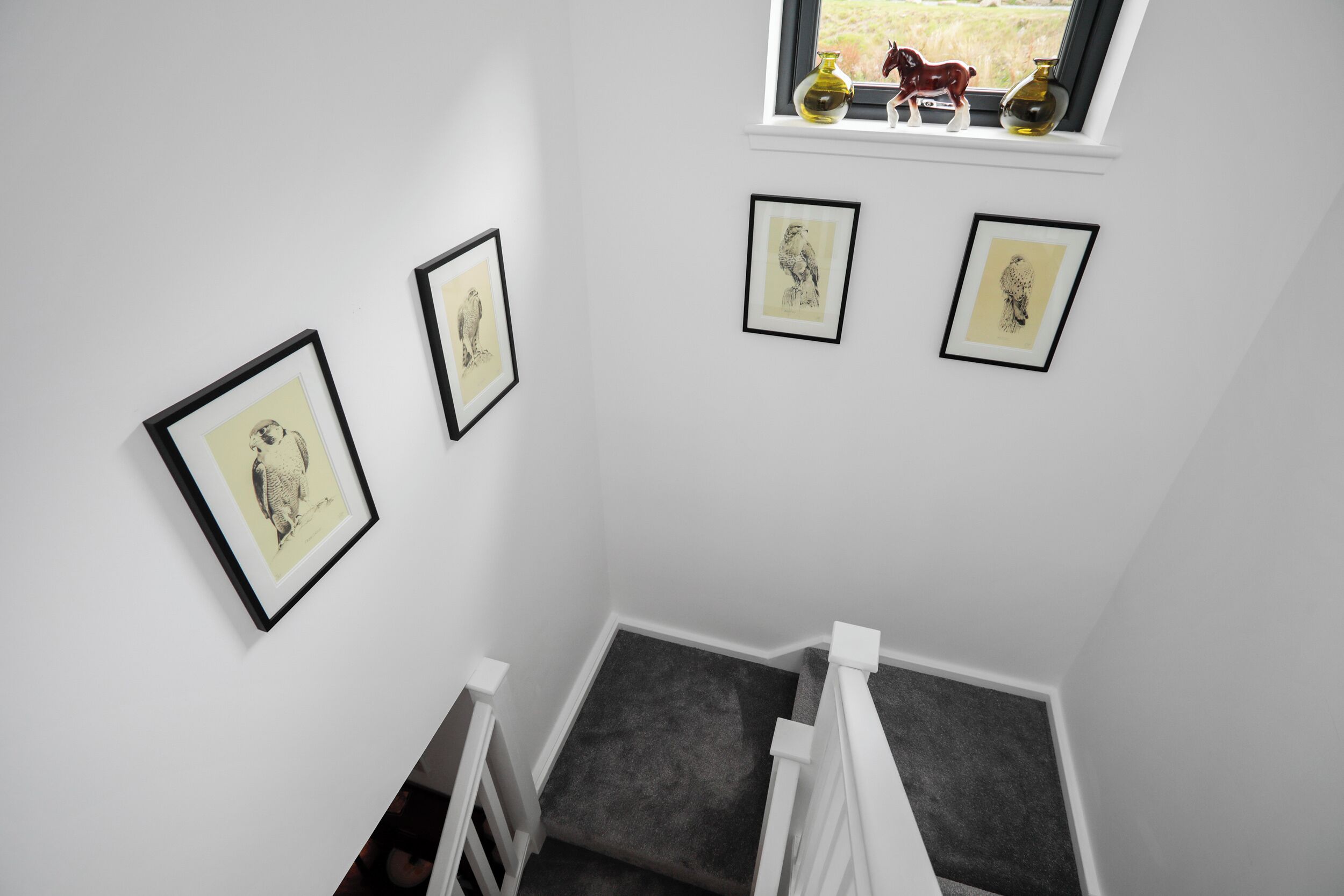 Una escalera con barandilla blanca, una ventana que muestra un caballo y un jarrón, y tres obras de arte enmarcadas en la pared.