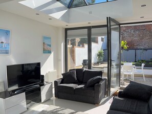 A modern living room with a flat-screen TV, grey sofa, and a glass door leading to an outdoor patio. - 23 Millhams Street (Christchurch)