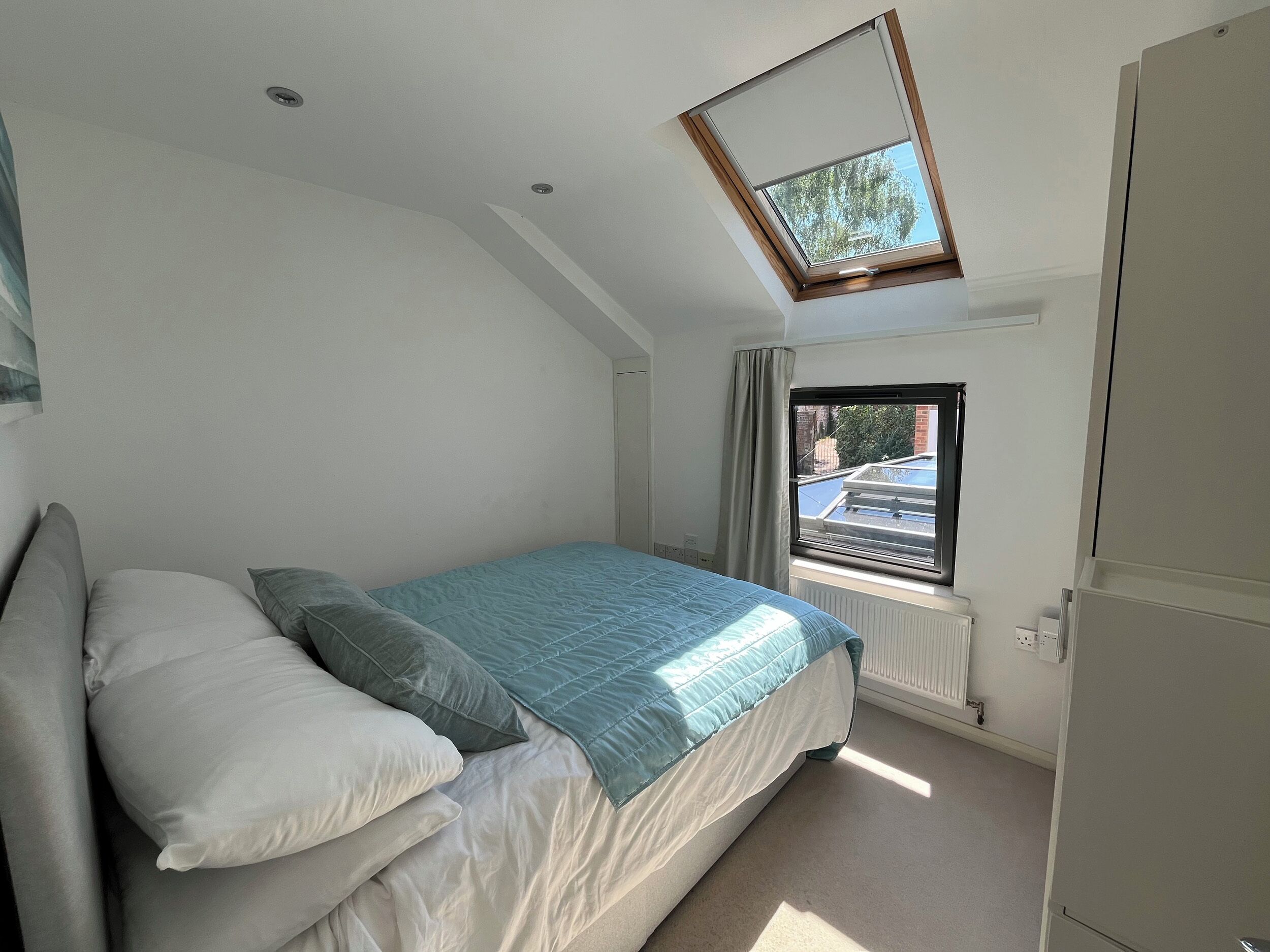 A bedroom with a skylight, a bed with pillows, a radiator, and a window with a view of the outside.