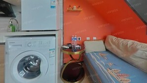 A compact hotel room with a washing machine, a small table with a water bottle, and a sofa with a floral pattern.