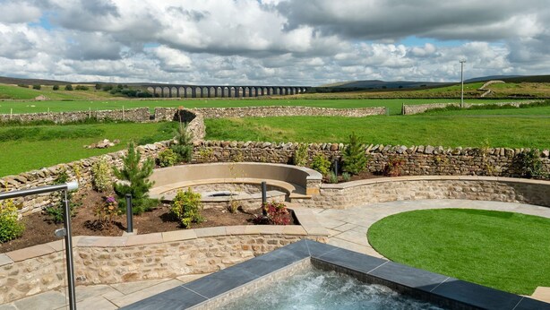 A hot tub area with stone walls and a stone pathway, surrounded by a well-maintained lawn and a stone wall. - Gunner Lodge (Carnforth)