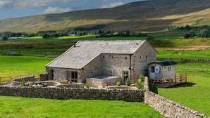 A stone farmhouse with a stone wall and a small wooden structure, surrounded by green fields and hills. - Gunner Lodge (Carnforth)