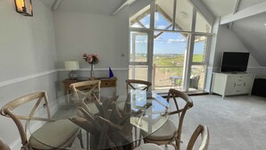 A dining area with a glass table and wooden chairs, a small desk with a lamp, a vase with flowers, and a view of the countryside through large windows. - 12 Thurlestone Rock (Kingsbridge)