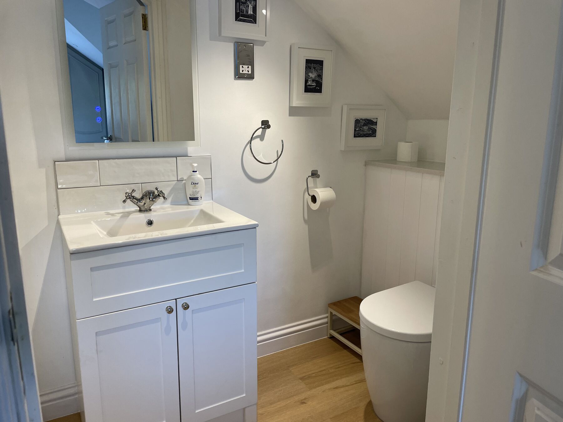 A bathroom with a white sink, mirror, and toilet. There are framed pictures on the wall and a small shelf with a roll of toilet paper.