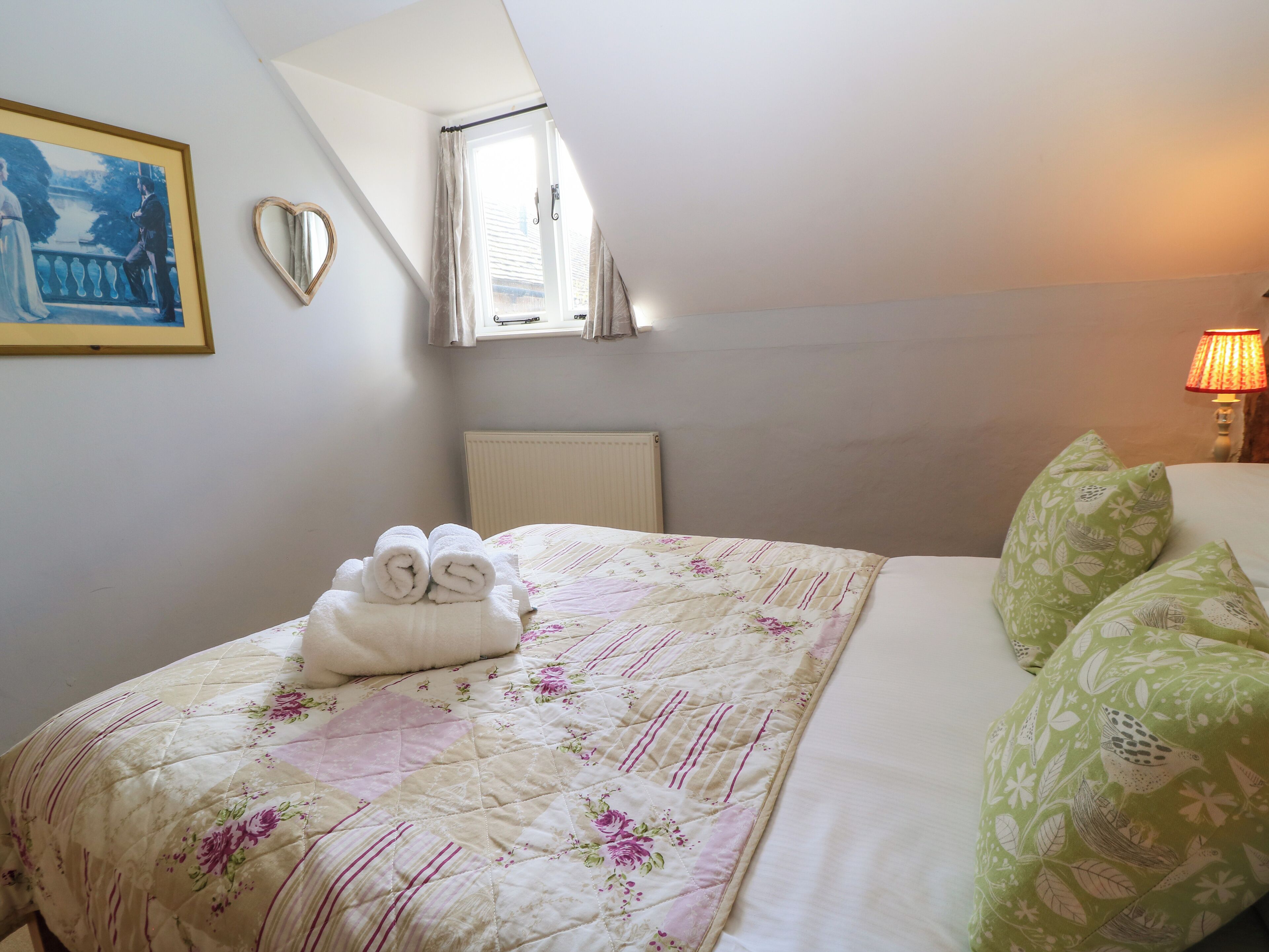 Una cama bien hecha con colcha floral, un par de toallas enrolladas, un espejo con forma de corazón y una lámpara en una mesita de noche.