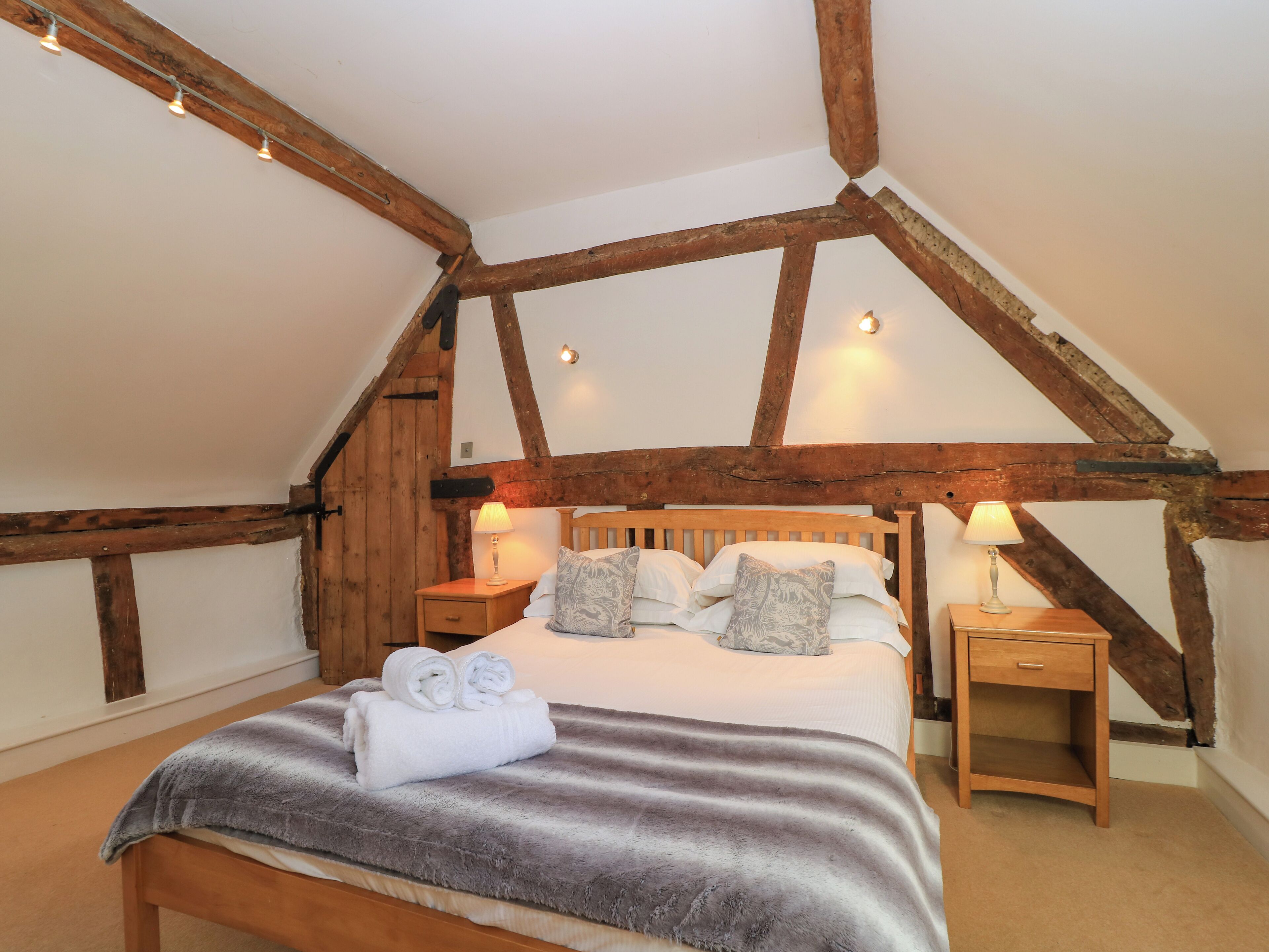 A bedroom with a sloped ceiling, exposed wooden beams, a wooden bed with multiple pillows, two bedside tables with lamps, and a rolled towel on the bed.