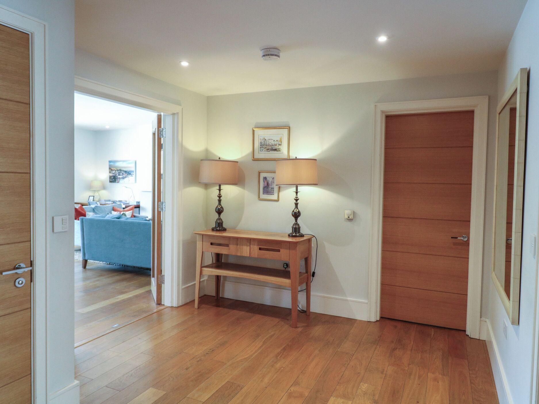 A hallway with wooden flooring, a desk with two lamps, a door, and a framed picture on the wall.