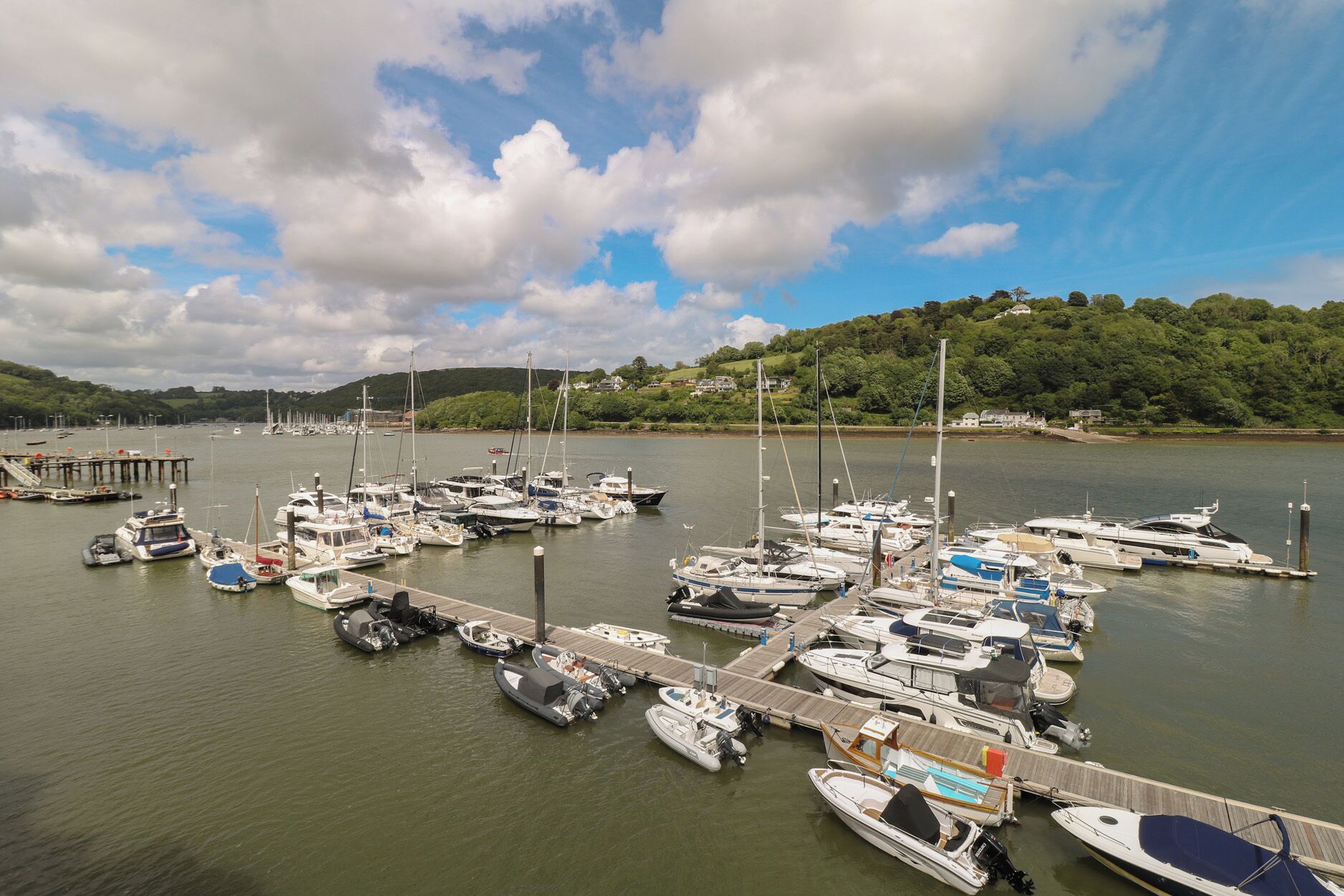 A marina with various boats docked, a wooden pier, and a backdrop of hills and trees.