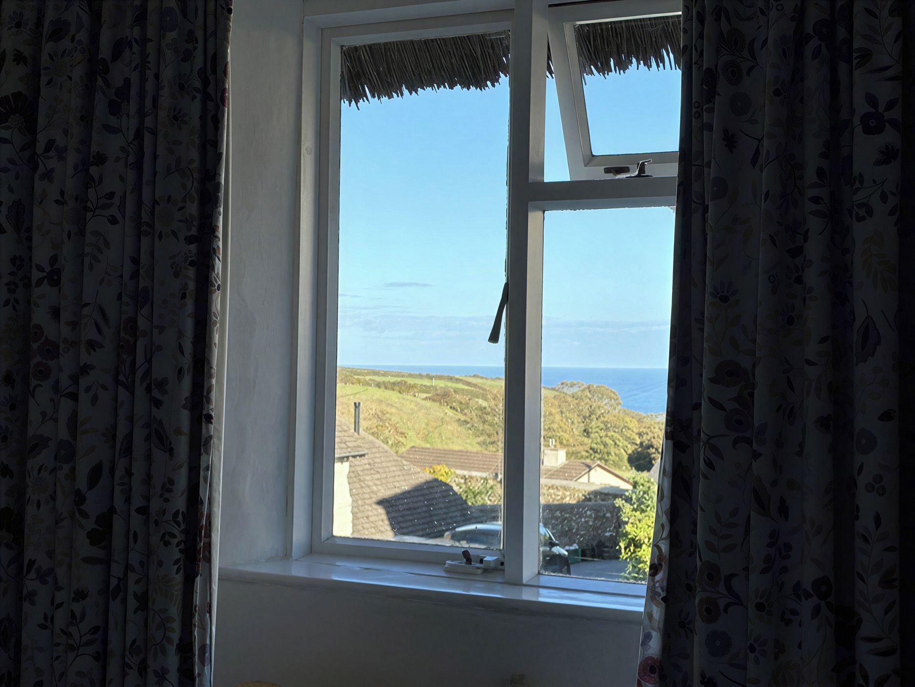 Une fenêtre donnant sur un paysage côtier, avec des rideaux et une petite table sur laquelle se trouve un appareil.