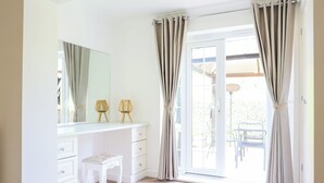 A bedroom with a white dresser, a small stool, a mirror, and beige curtains. - Howard Cottage (Evesham)