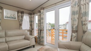 Ein Wohnzimmer mit einem beigen Sofa, einem kleinen Tisch und Blick auf eine Holzterrasse durch eine Glastür.
