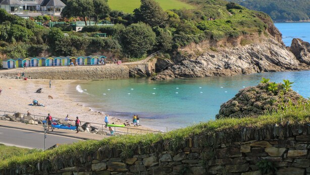 A coastal beach with a stone wall, a row of colorful beach huts, and a grassy hill with houses in the background. - 2 The Dunes (Falmouth)
