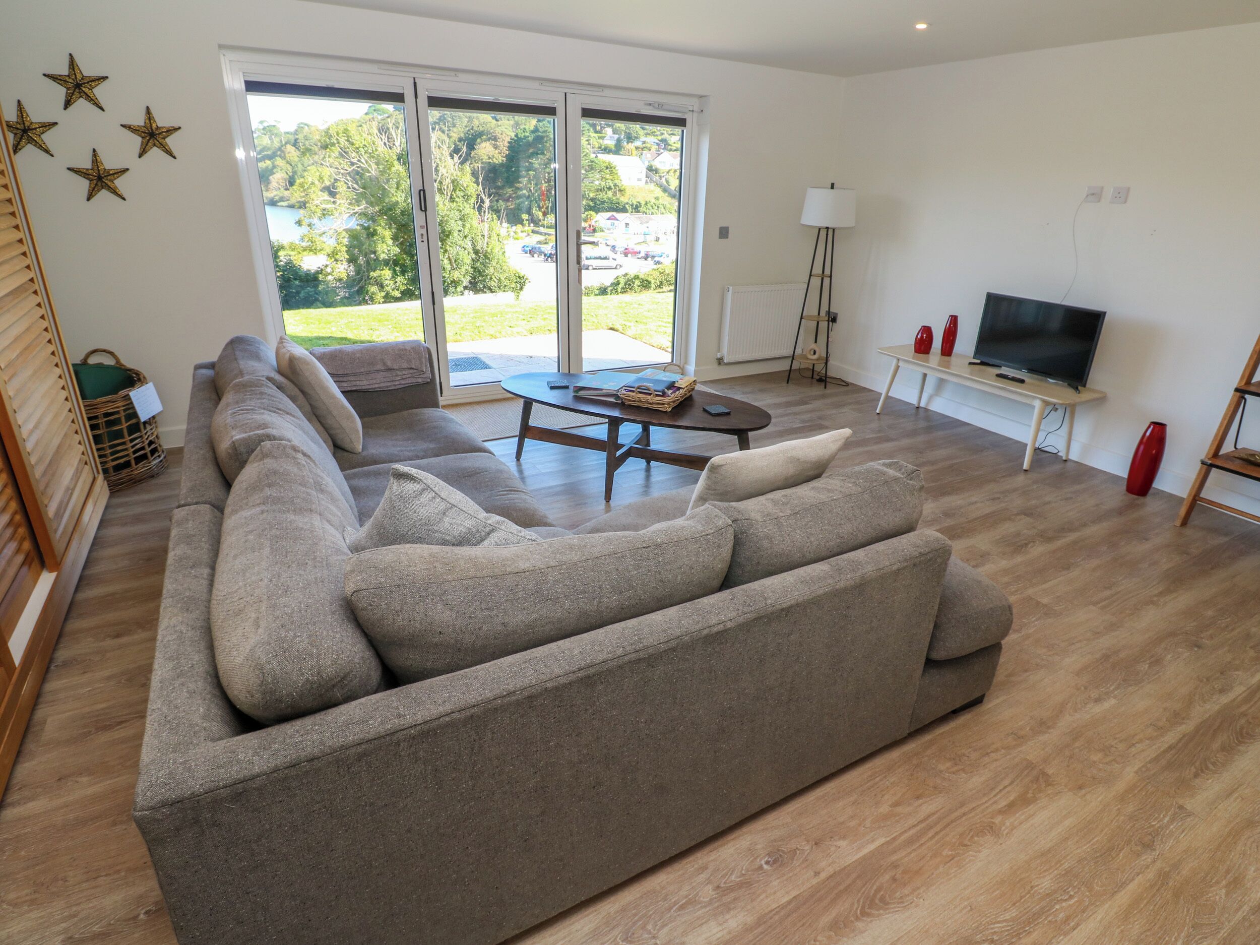A living room with a large grey sectional sofa, a coffee table, a TV on a stand, and a view of a green outdoor area through large sliding doors.