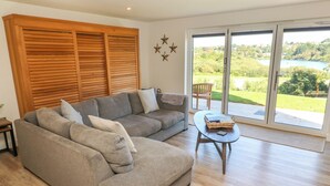 A modern living room with a grey sectional sofa, a small wooden coffee table, and a view of a lake through large sliding glass doors. - 2 The Dunes (Falmouth)