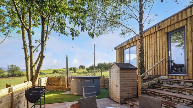A patio with a table and chairs, a barbecue grill, a hot tub, and a wooden building with a window. - Orchard Lodge (Corwen)