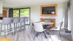 A modern dining area with a round table and bar stools, a flat-screen TV mounted on the wall, and a view of the outdoors through large windows. - Orchard Lodge (Corwen)