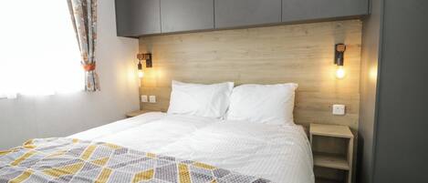 Une chambre moderne avec une tête de lit en bois, un lit recouvert de draps blancs, un plaid à motifs géométriques et des lampes de chevet.