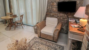 A living room with a stone wall, a flat-screen TV mounted above a wooden side table, a wicker armchair, a dining table with chairs, and a large window with blinds. - The Coquet Nest (Morpeth)
