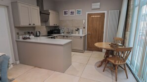 A modern kitchen with a central island, a dining area with a wooden table and chairs, and a view through large windows. - The Coquet Nest (Morpeth)